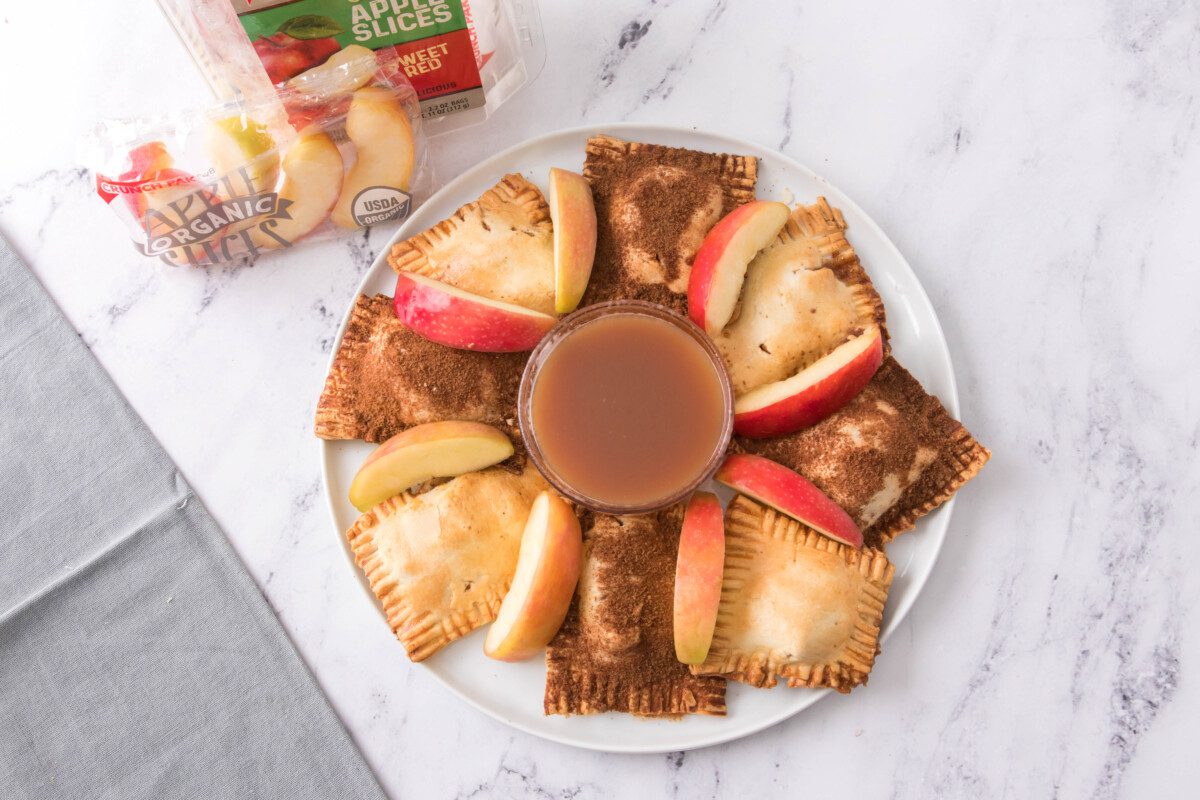 horizontal overhead shot of air fryer apple hand pies on a white plate with caramel sauce for dipping and sliced apples