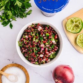 overhead vertical shot of cilantro, juicy gems arils, squeezed lemon on cutting board, red onion and pomegranate salsa in a white bowl