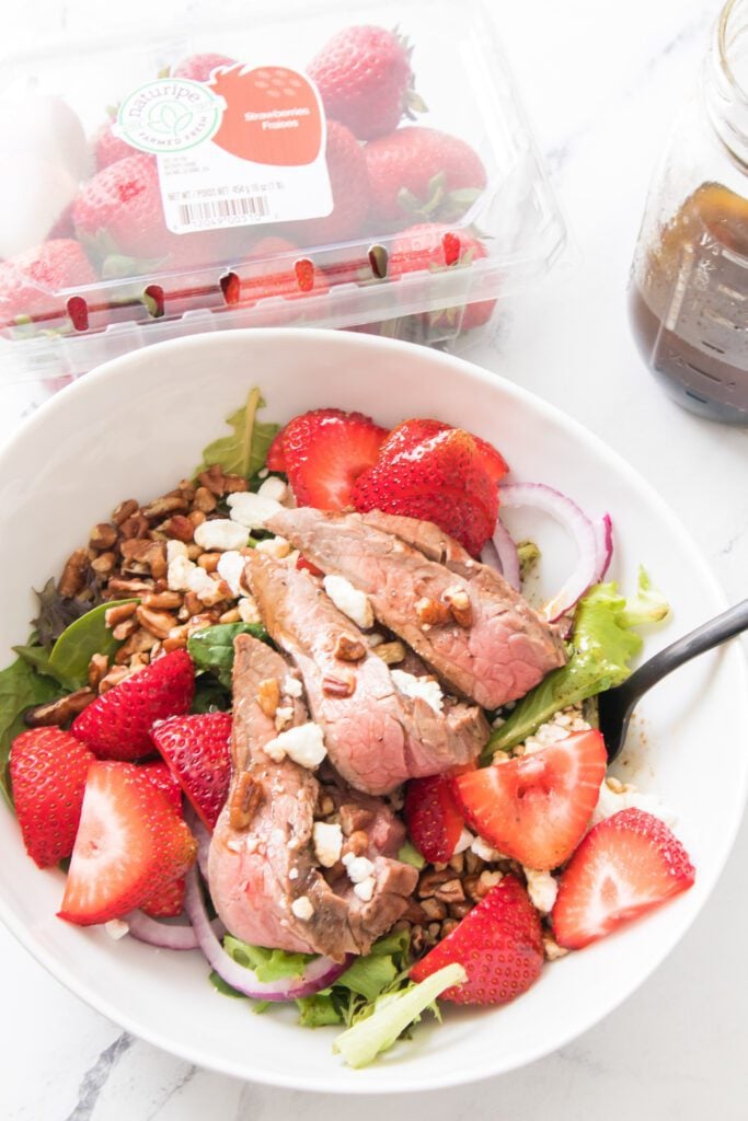 vertical image of strawberry salad with steak in white bowl with fork