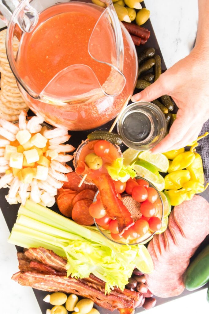 overhead shot of bloody mary in glass pitcher with hand reaching for vodka on a bloody mary charcuterie board