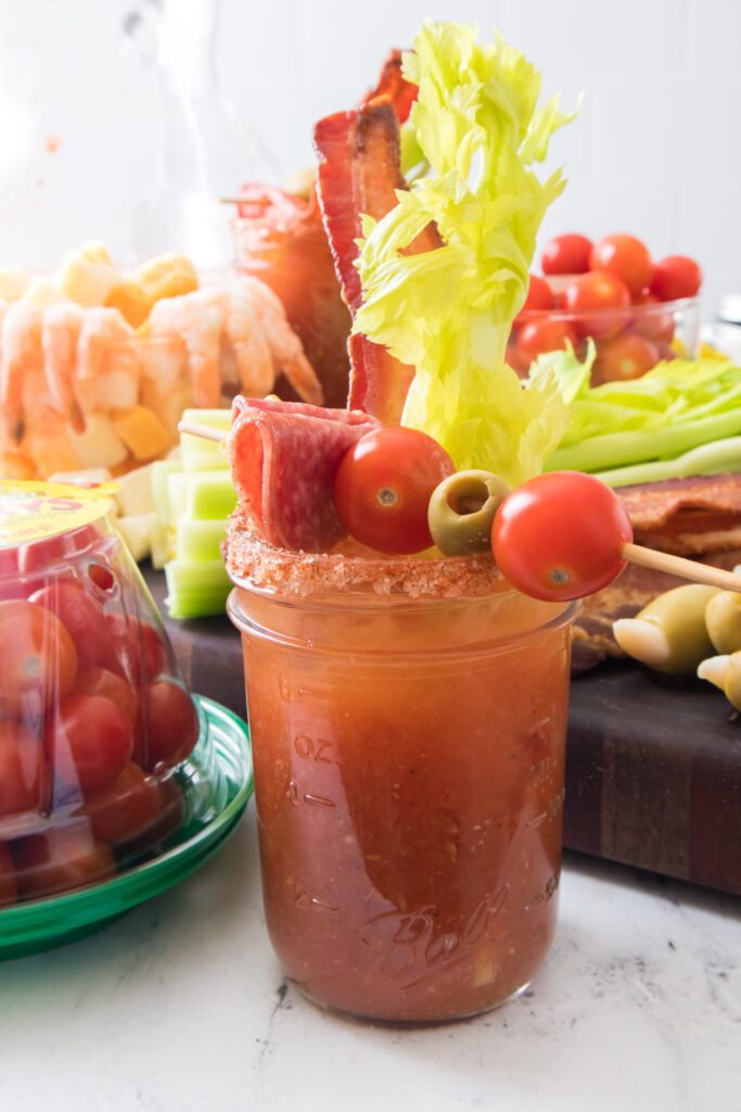 vertical shot of bloody mary in mason jar with tomatoes and bloody mary chartcuterie board in background