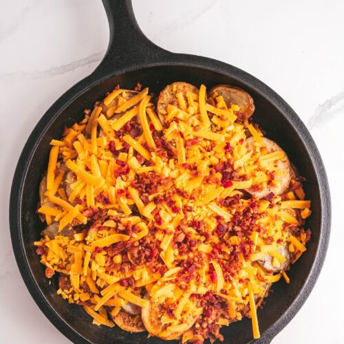 Flat lay photo of a skillet with baked potato slices topped with shredded sharp cheddar cheese and cooked chopped bacon