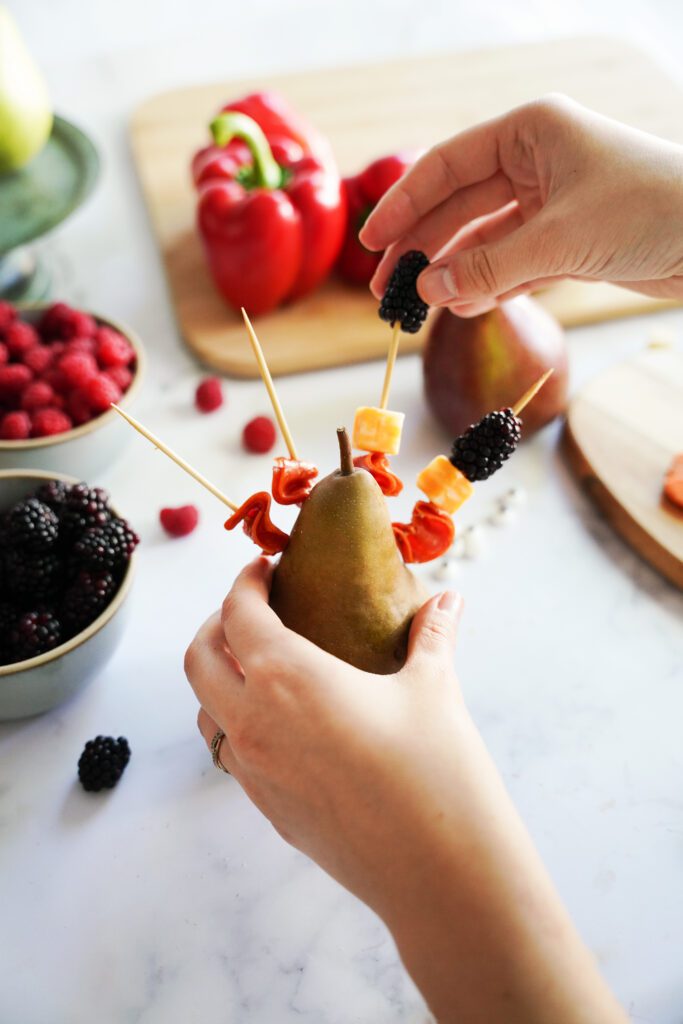 skewers inserted into a pear with pepperoni slices, cubed cheese and blackberries