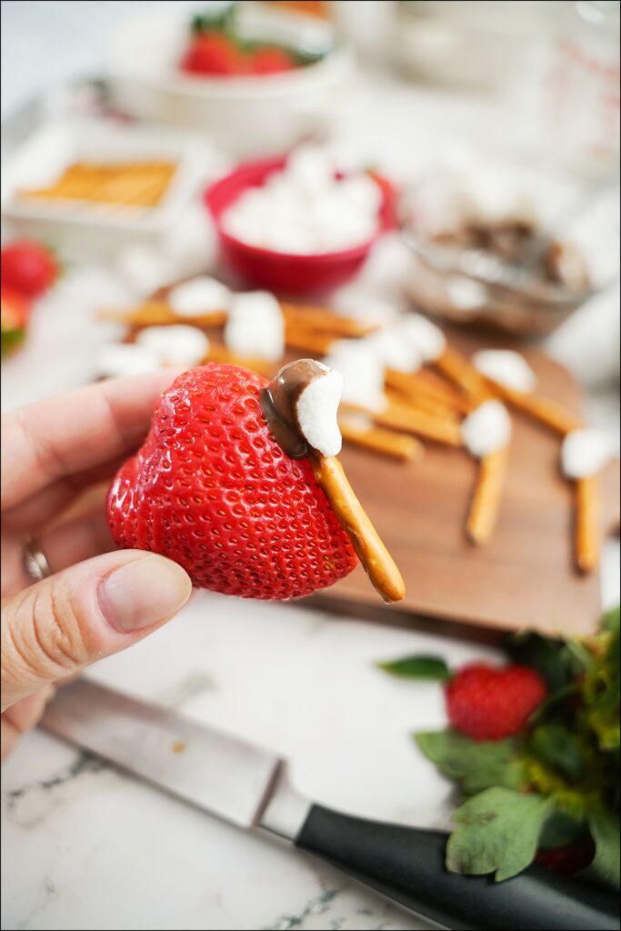 hand holding strawberry with melted chocolate, mini marshmallow attached to a pretzel stick