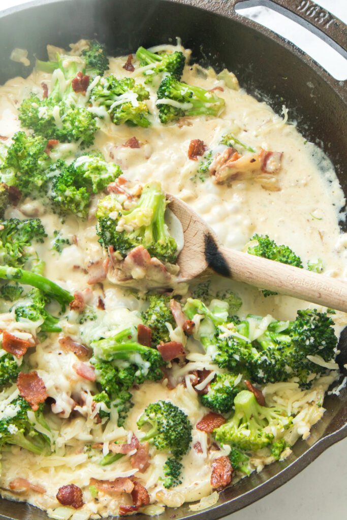 vertical close up shot of broccoli in creamy garlic sauce with cheese and bacon on wooden spoon