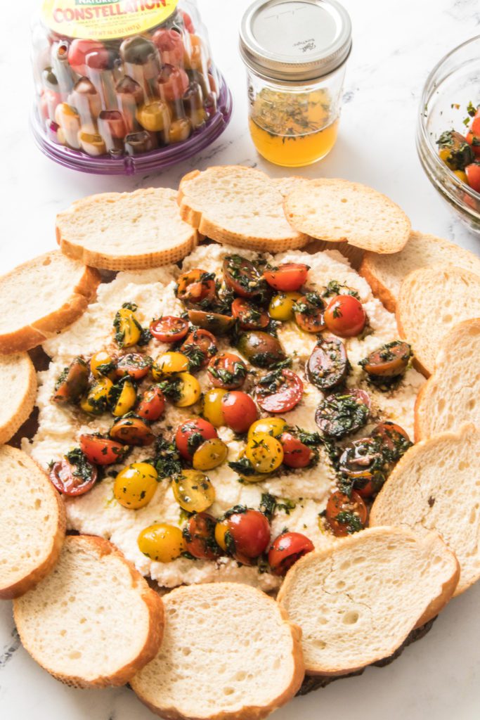 close up vertical shot of bruschetta board with ricotta, fresh tomatoes, herbs, lemon honey dressing in mason jar and tub of naturesweet constellation tomatoes
