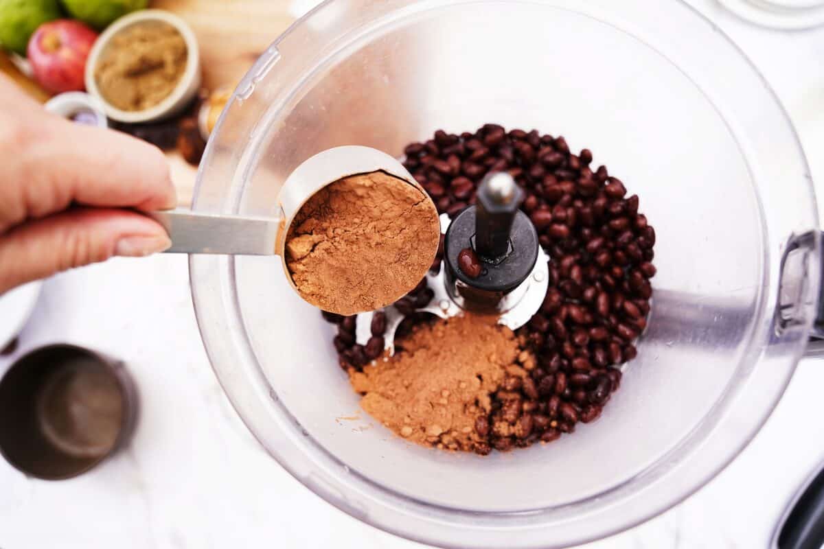 overhead shot of food processor with black beans and cocoa powder being placed in measuring cup