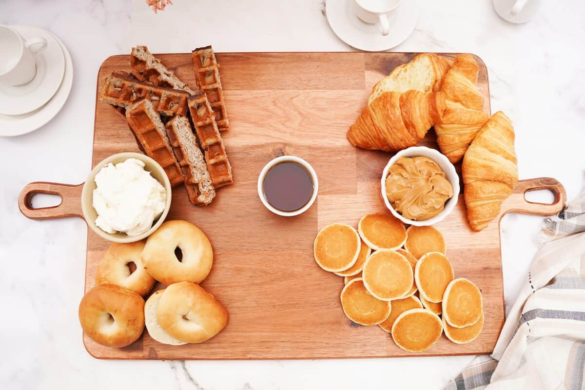 overhead shot of board with mini pancakes, waffles, bagels and croissants with whipped cream, syrup and peanut butter in bowls