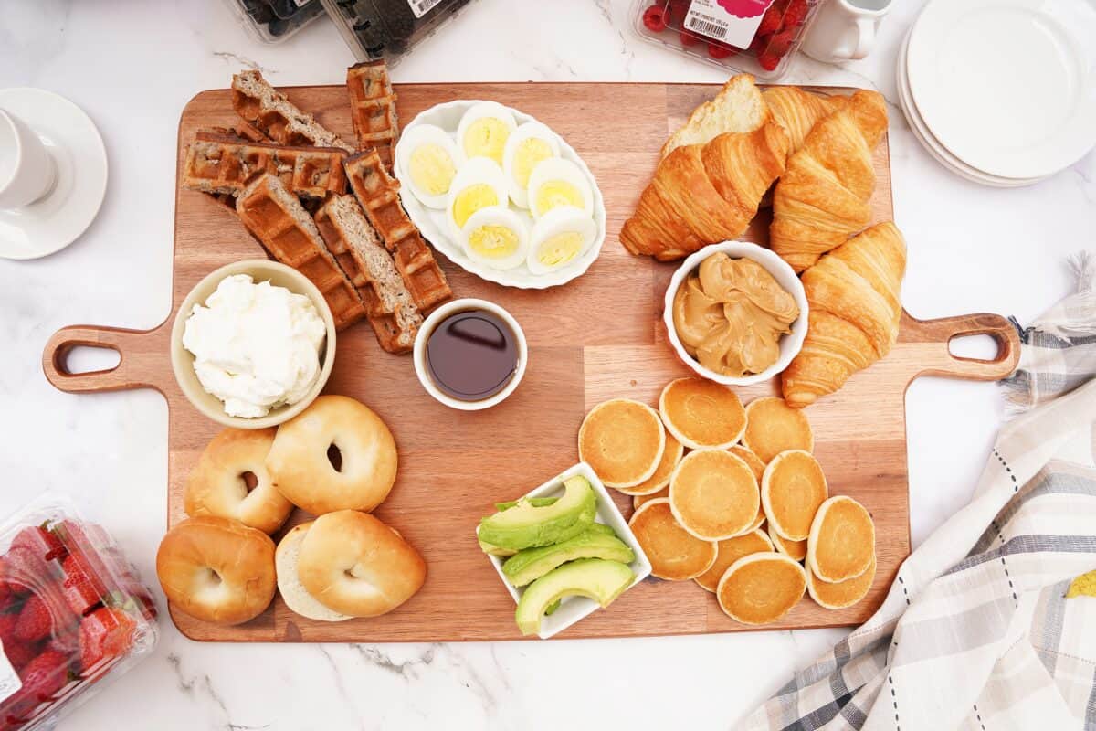 overhead shot of board with mini pancakes, waffles, bagels and croissants with whipped cream, syrup and peanut butter in bowls with avocado slices and sliced hard boiled eggs on platter