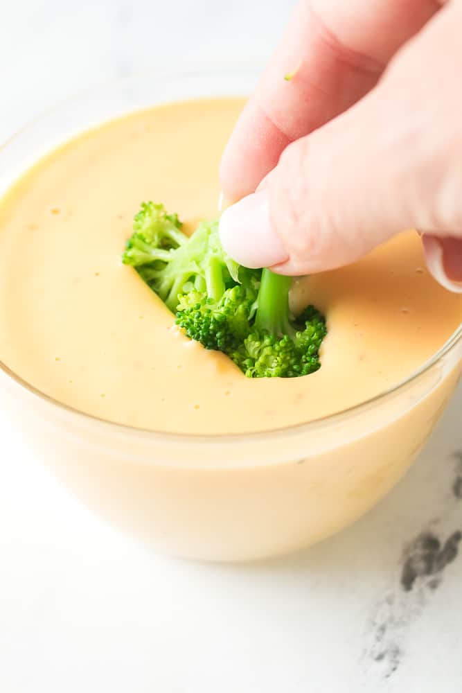 hand dipping broccoli into bowl of cheese sauce