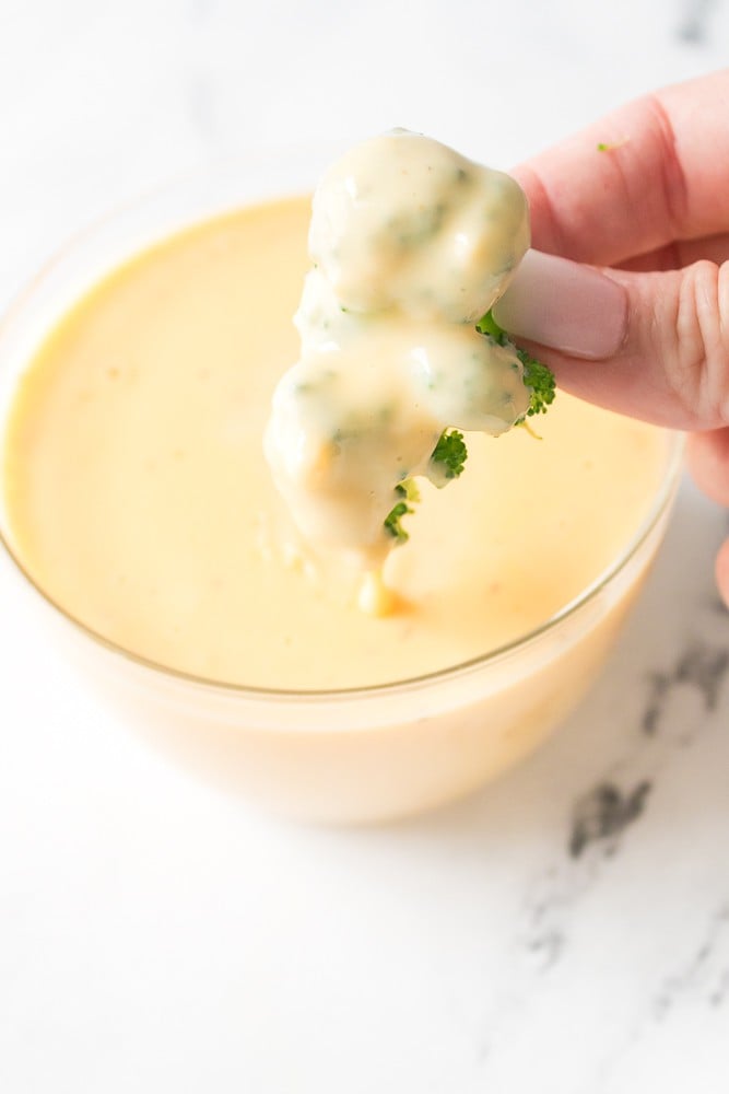 hand holding cheese covered broccoli floret over bowl of broccoli cheese sauce