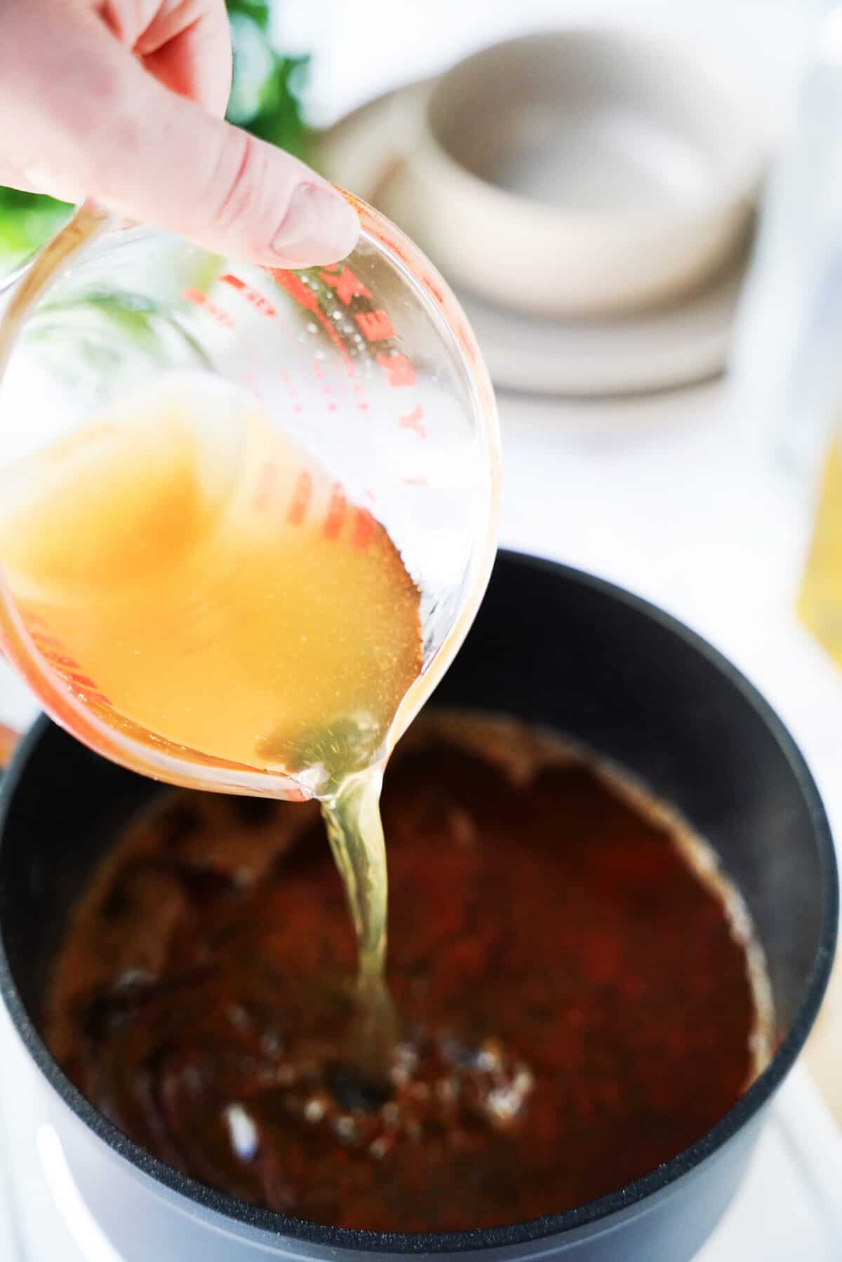 measuring cup pouring vegetable broth into sauce pan with oil and flour