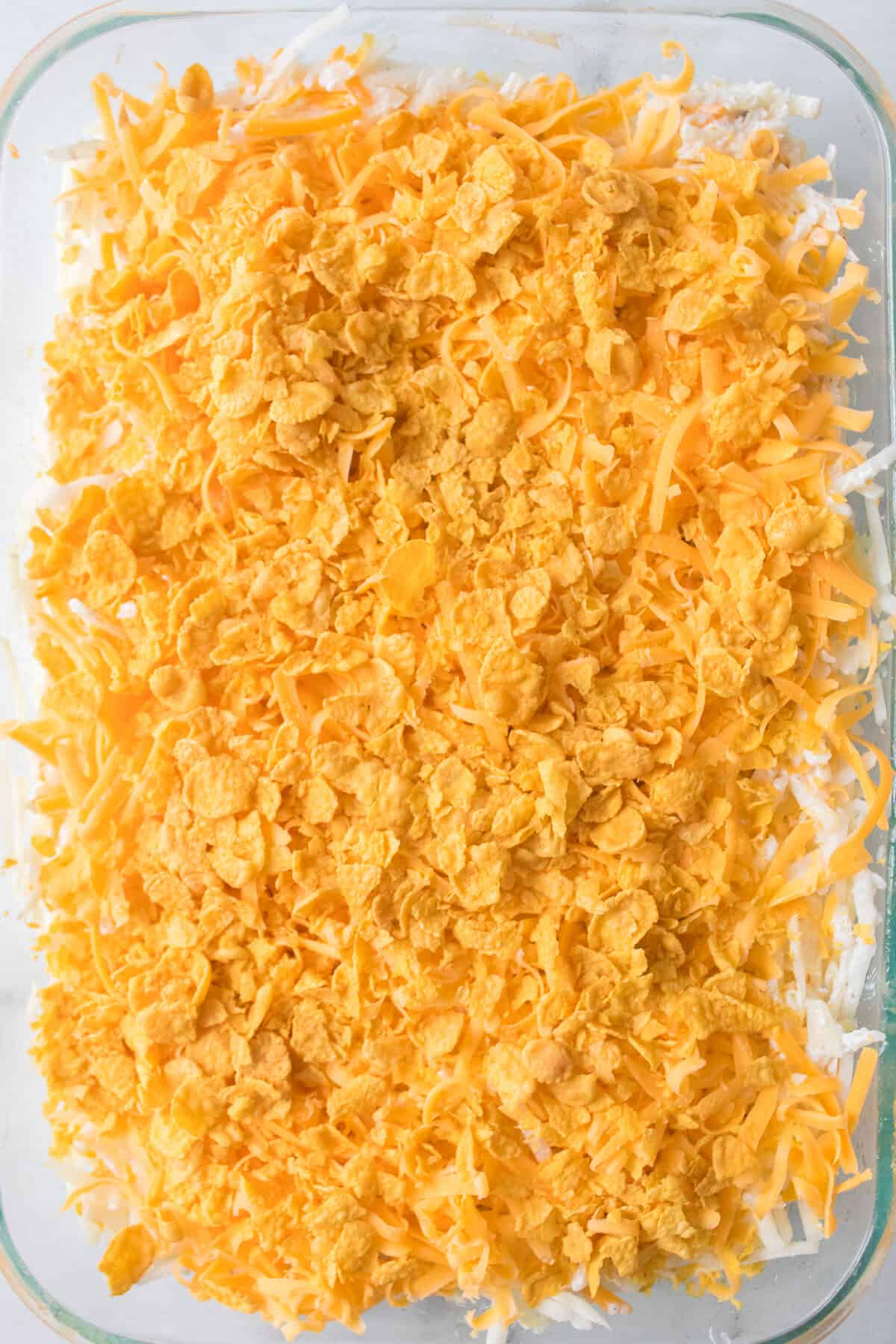 unbaked funeral potatoes topped with cheese and corn flakes in casserole dish