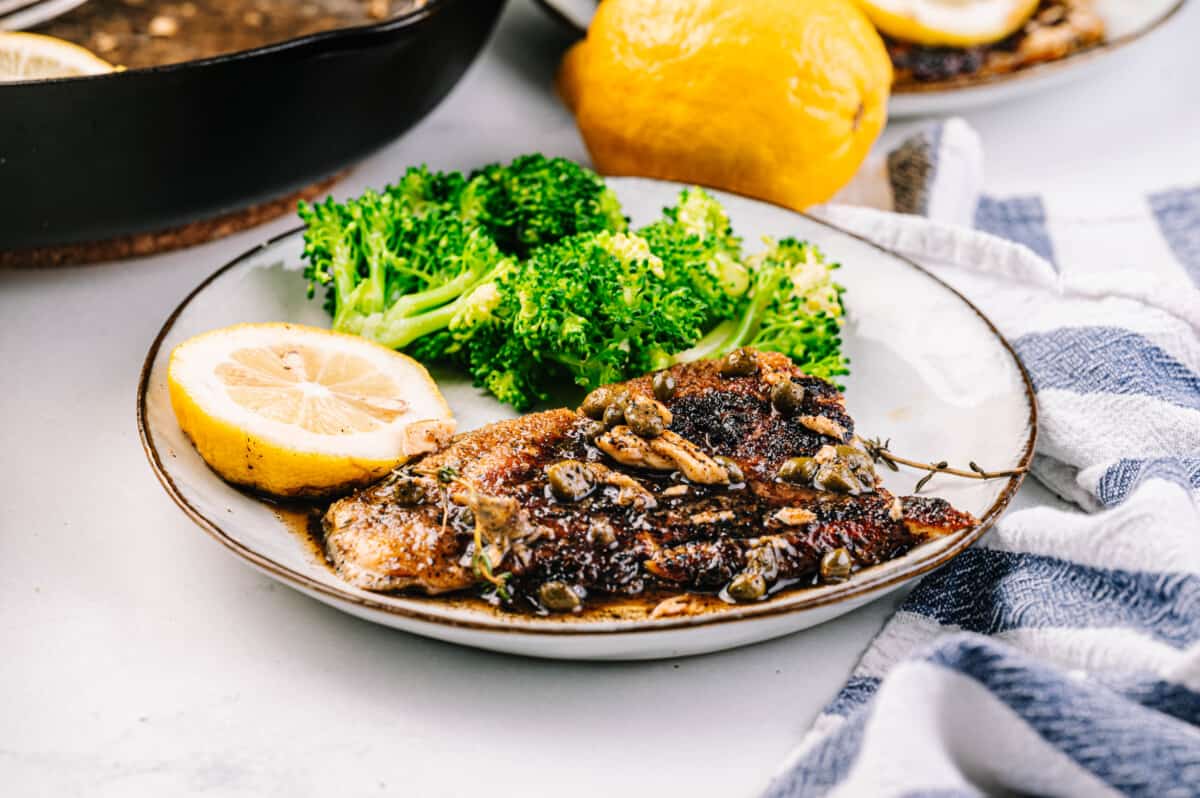 Fish piccata with lemon caper sauce with a side of broccoli on a plate.
