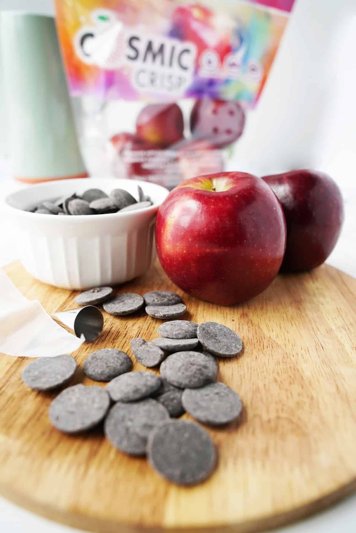 Chocolate melts, apples, wax paper, and a piping tip on a wooden cutting board.