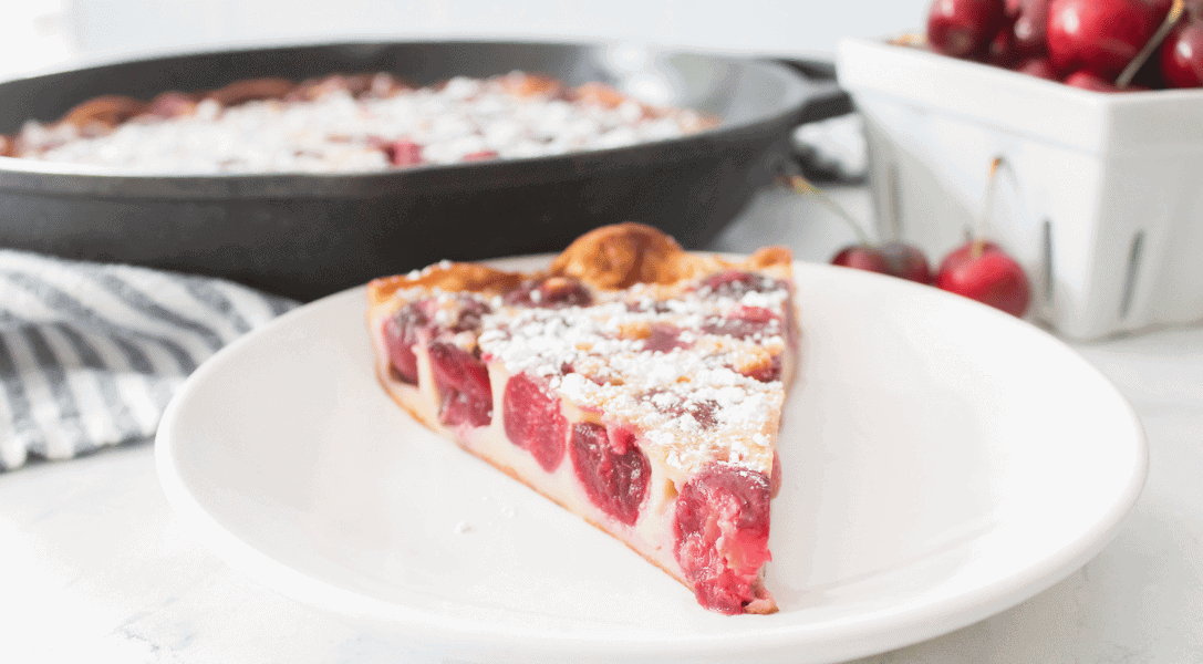 Slice of Cherry Clafoutis on white plate with rest of Cherry Clafoutis in pan