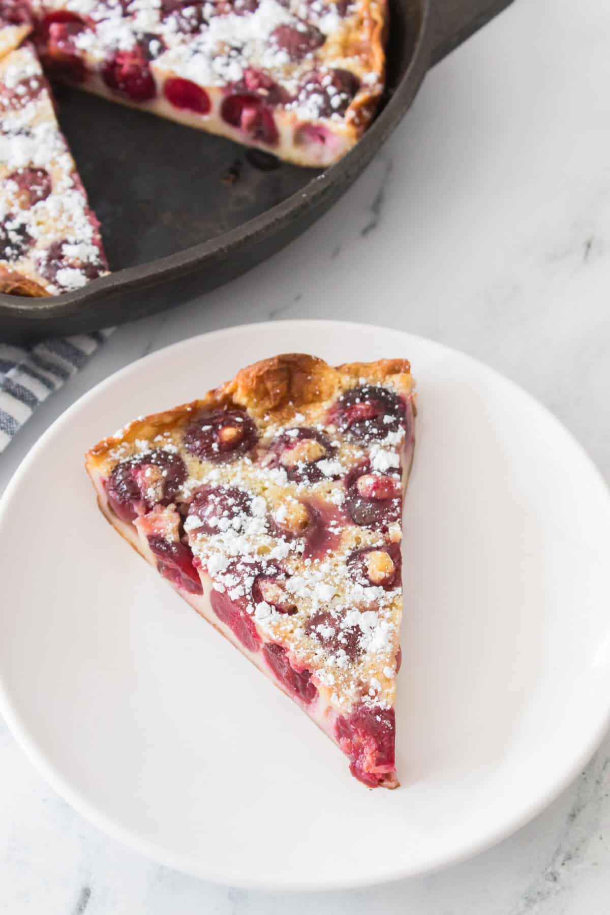 vertical overhead shot of a slice of cherry clafoutis on a white plate dusted with powdered sugar
