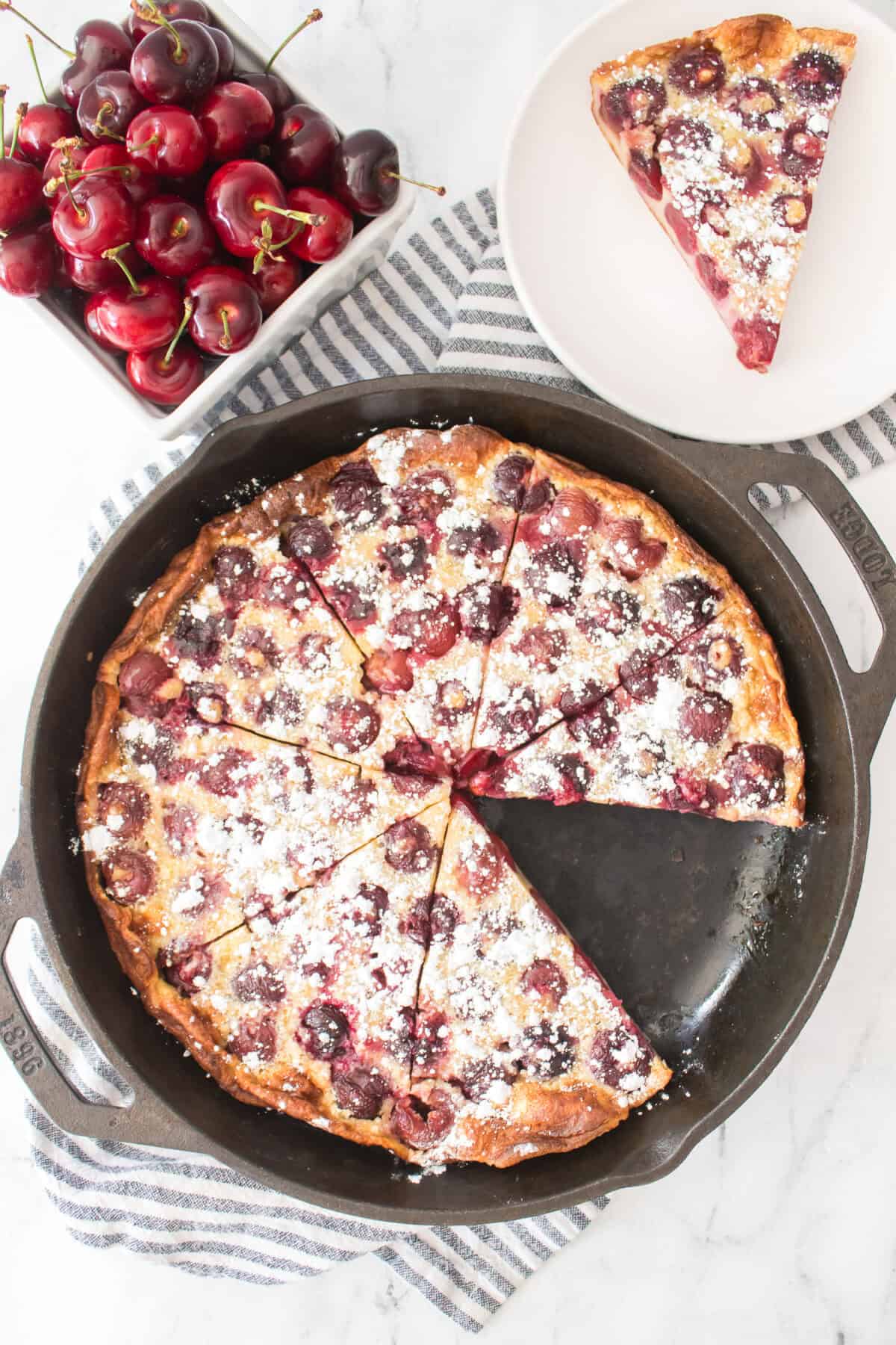 overhead vertical shot of cherry clafoutis baked in cast iron skillet with a slice missing