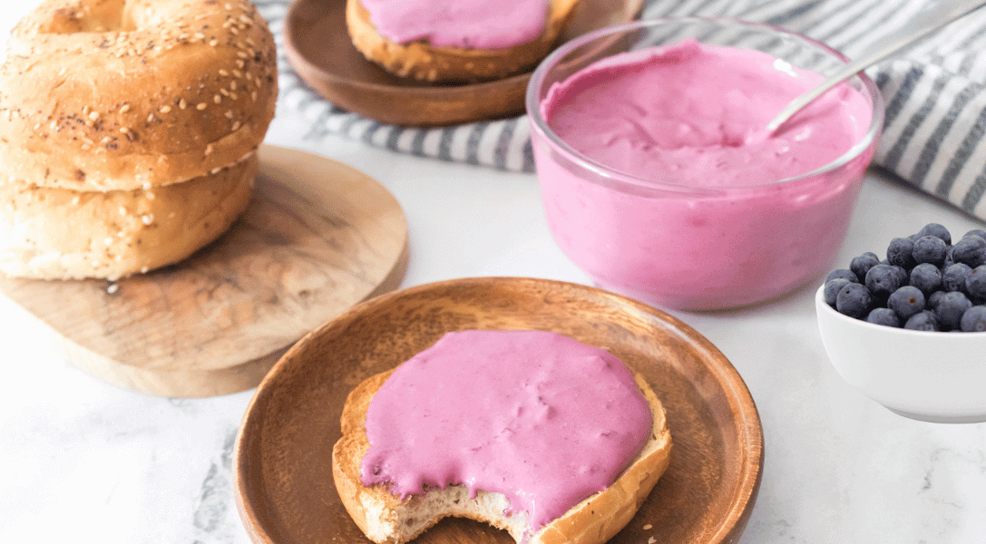 Blueberry Cream Cheese on bagel surrounded by more bagels, blueberries in a bowl and cream cheese in a bowl