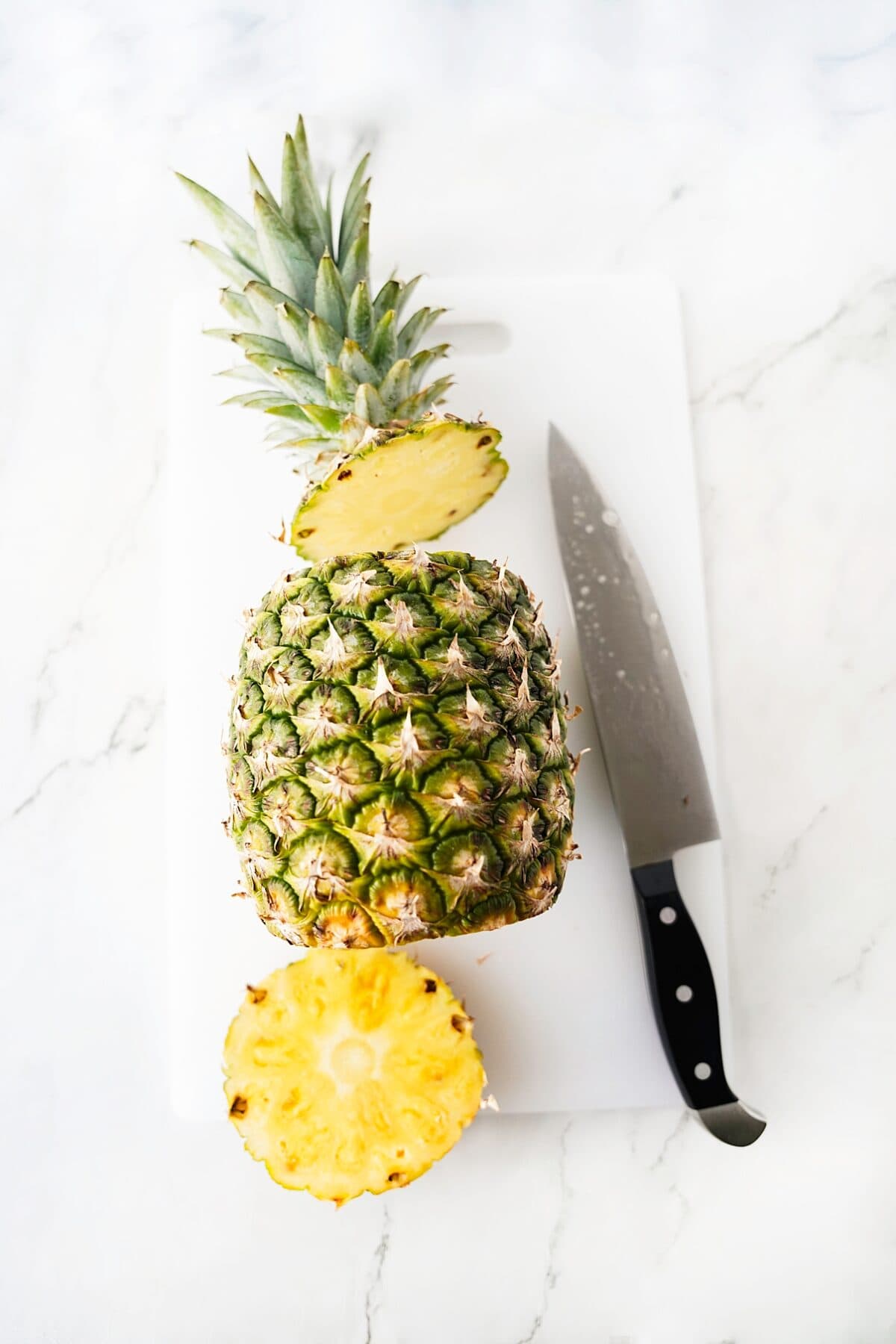 overhead shot of pineapple with top and bottom cut off with knife on cutting board