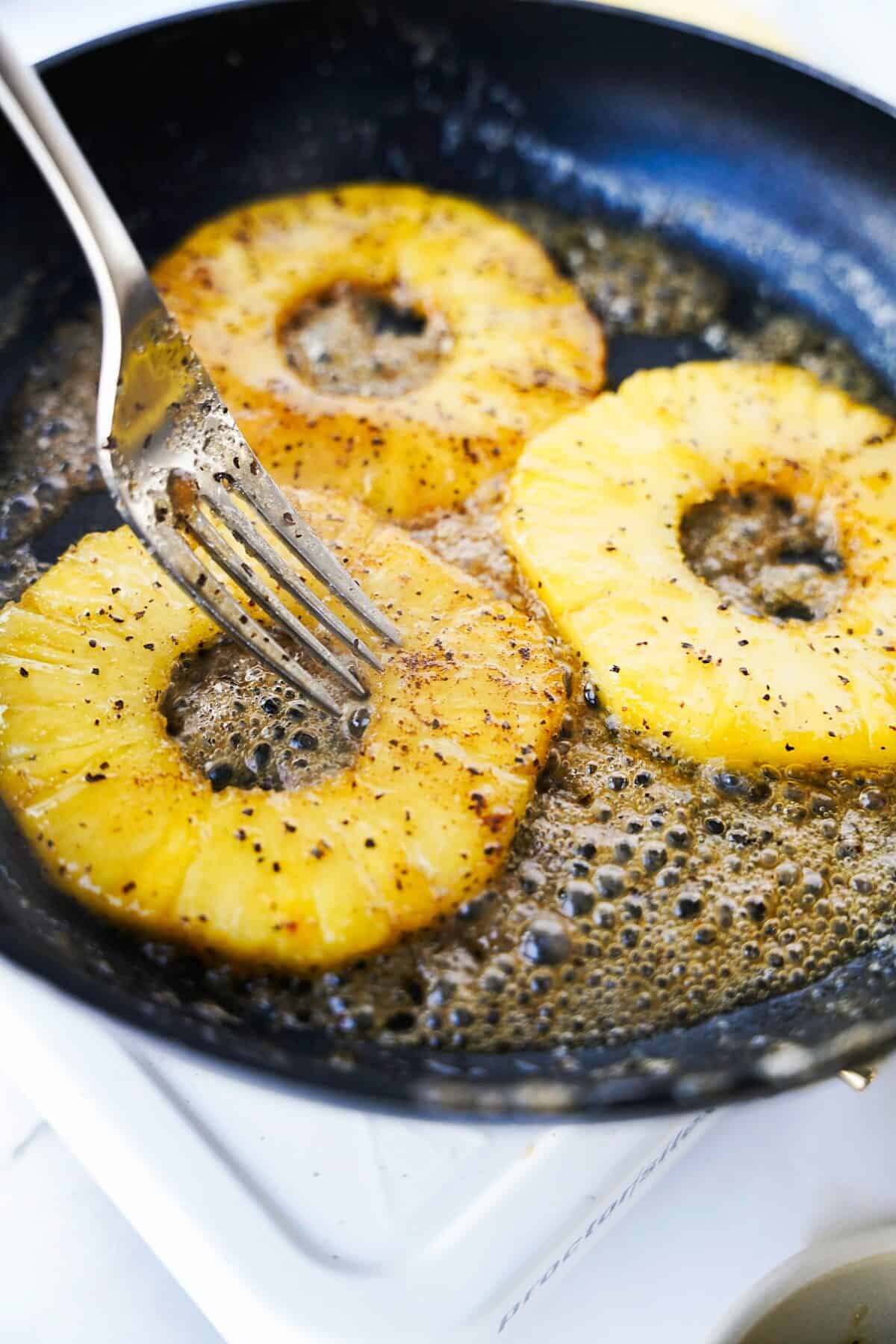 cast iron skillet cooking spicy pineapple slices in butter with fork preparing to flip over to caramelize on the other side