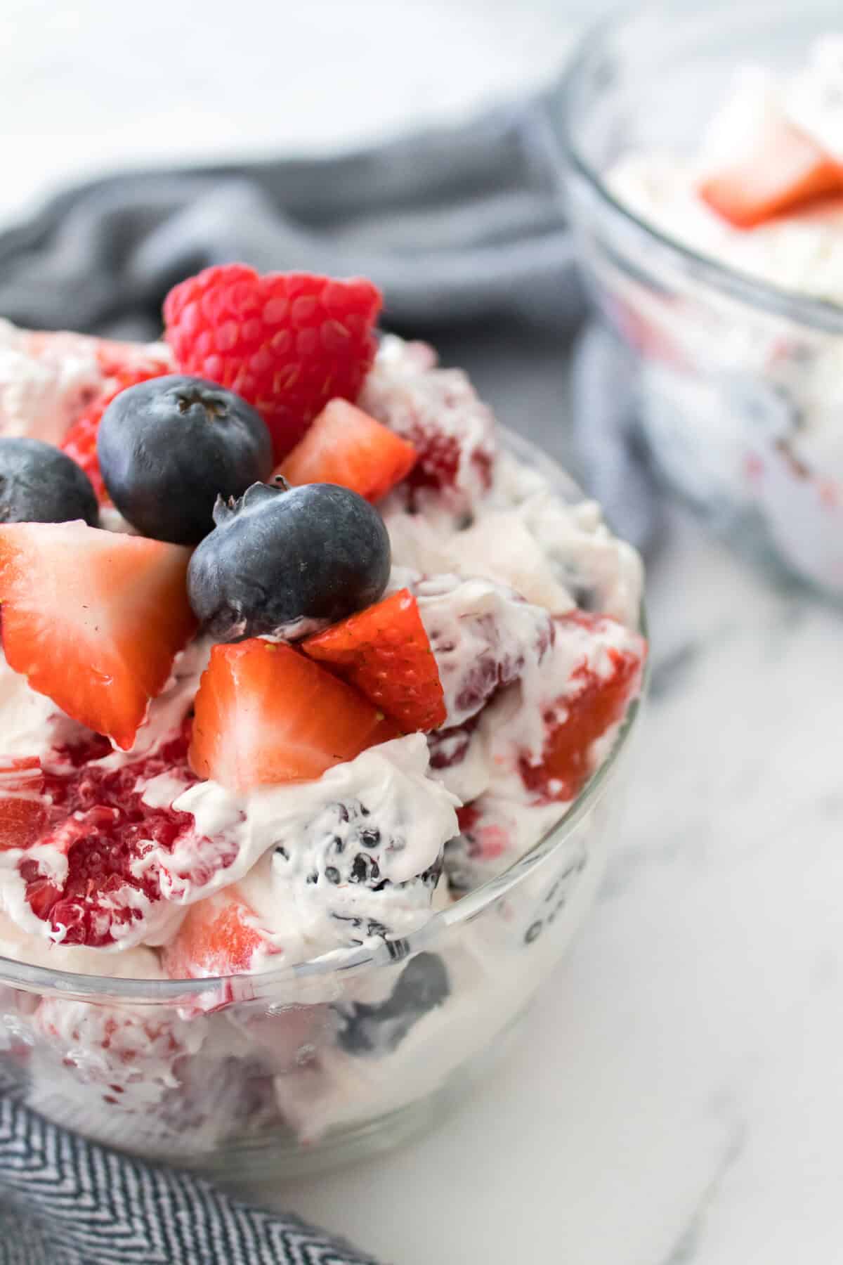 close up vertical shot of summertime cheesecake fruit salad in glass serving bowl topped with blueberries, raspberries and strawberries with bowl in background of cheesecake fruit salad