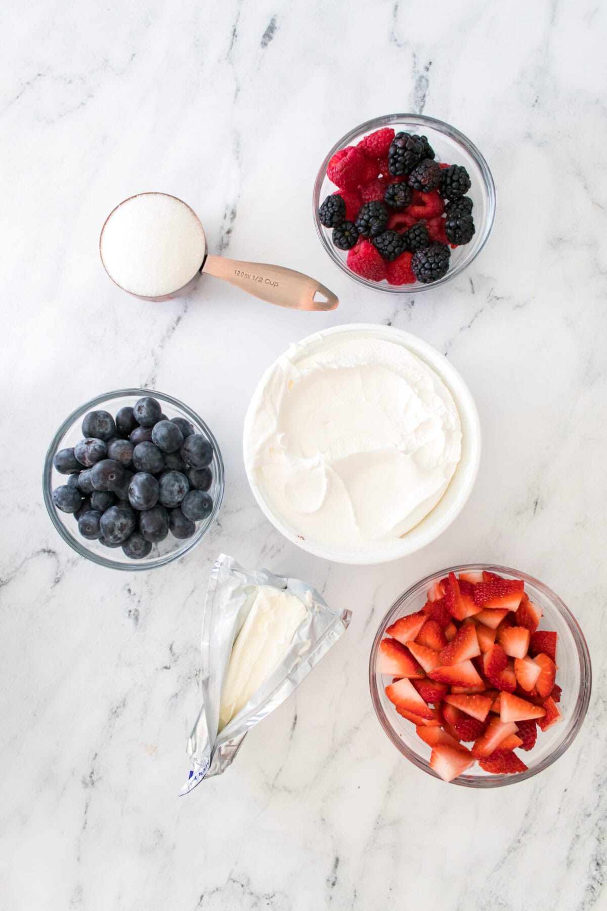 overhead shot of ingredients needed for summertime cheesecake fruit salad: blackberries and raspberries in glass dish, copper 1/2 c measuring cup with sweetener, glass bowl of blueberries, tub of whipped topping, 8 oz of cream cheese and glass bowl of quartered strawberries