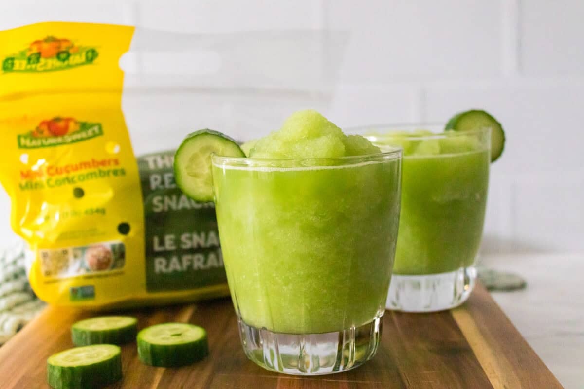 head on shot of cucumber slushies garnished with slice of cucumber sitting on cutting board with bag of naturesweet mini cucumbers