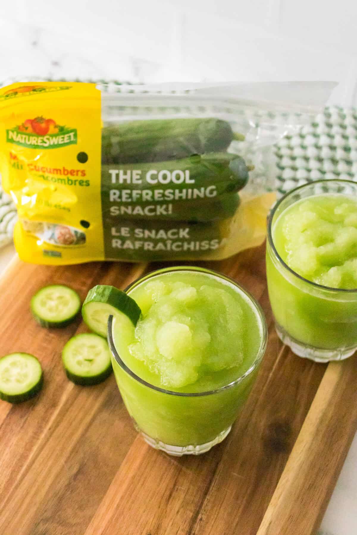 45 degree shot of cucumber slushies in glasses garnished with slice of cucumber, bag of naturesweet mini cucumbers on cutting board