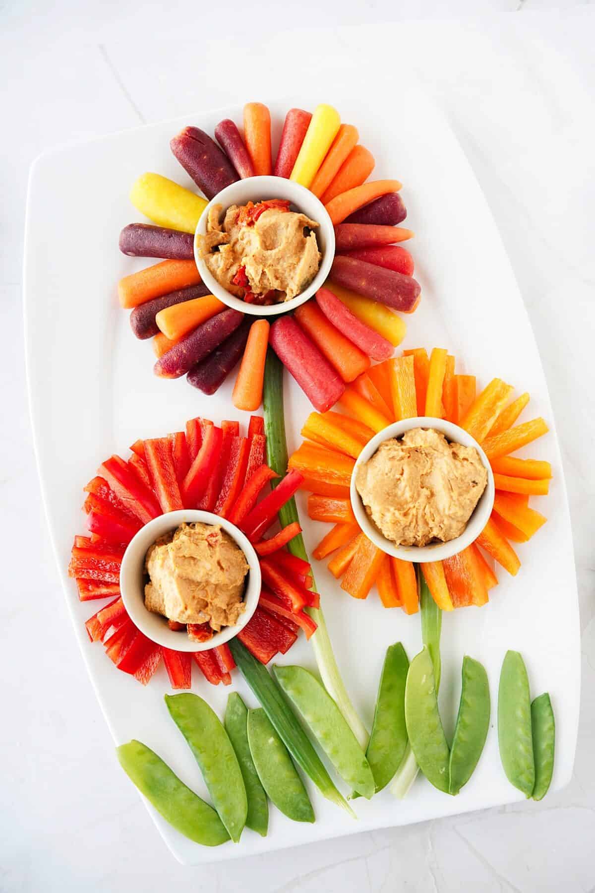 overhead shot of colored baby carrots, red and orange bell peppers with bowls of hummus in the center to create flowers with stems of green onions and snap peas as grass on the bottom