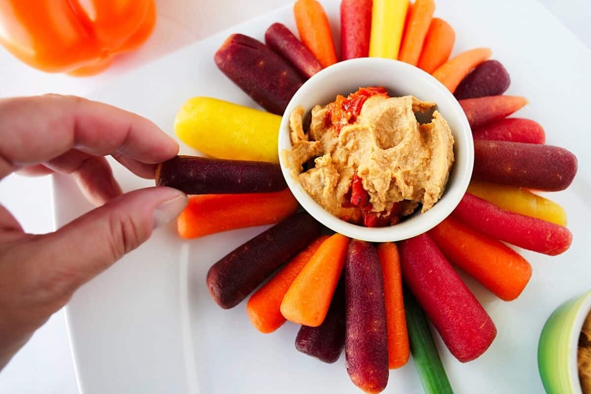 close up of hand grabbing a colored baby carrot to dip in bowl of hummus