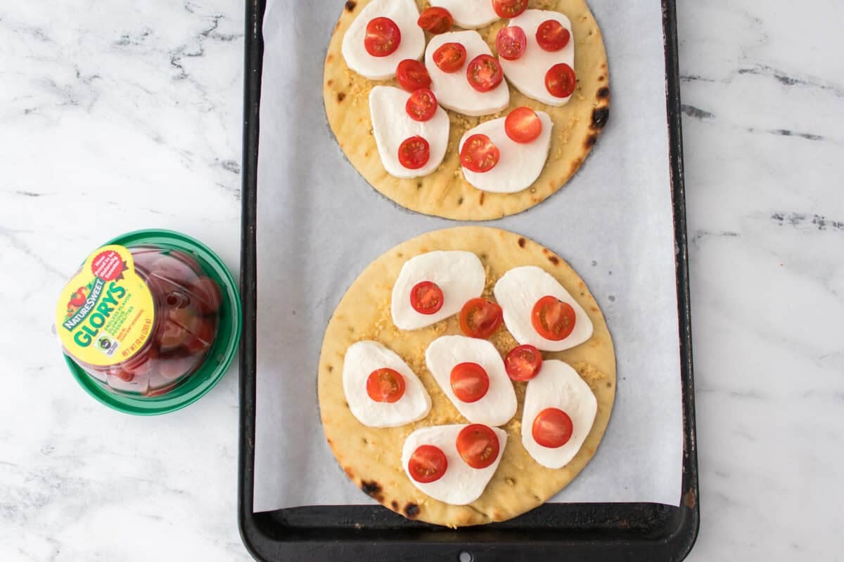 overhead shot of baking sheet lined with parchment paper with two flatbreads, drizzled with olive oil, minced garlic, mozzarella cheese slices and cherry tomatoes before baking