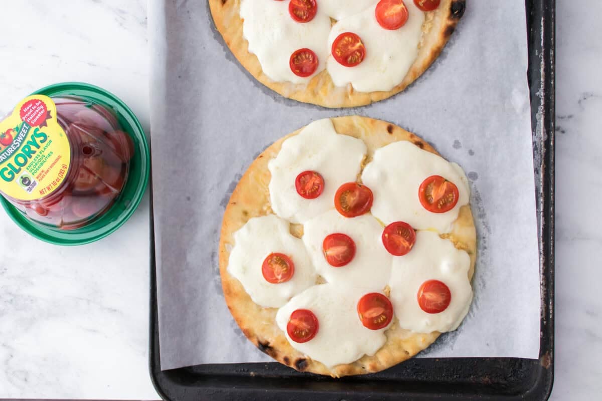 overhead shot of baking sheet lined with parchment paper with two flatbreads, drizzled with olive oil, minced garlic, mozzarella cheese slices and cherry tomatoes after baking