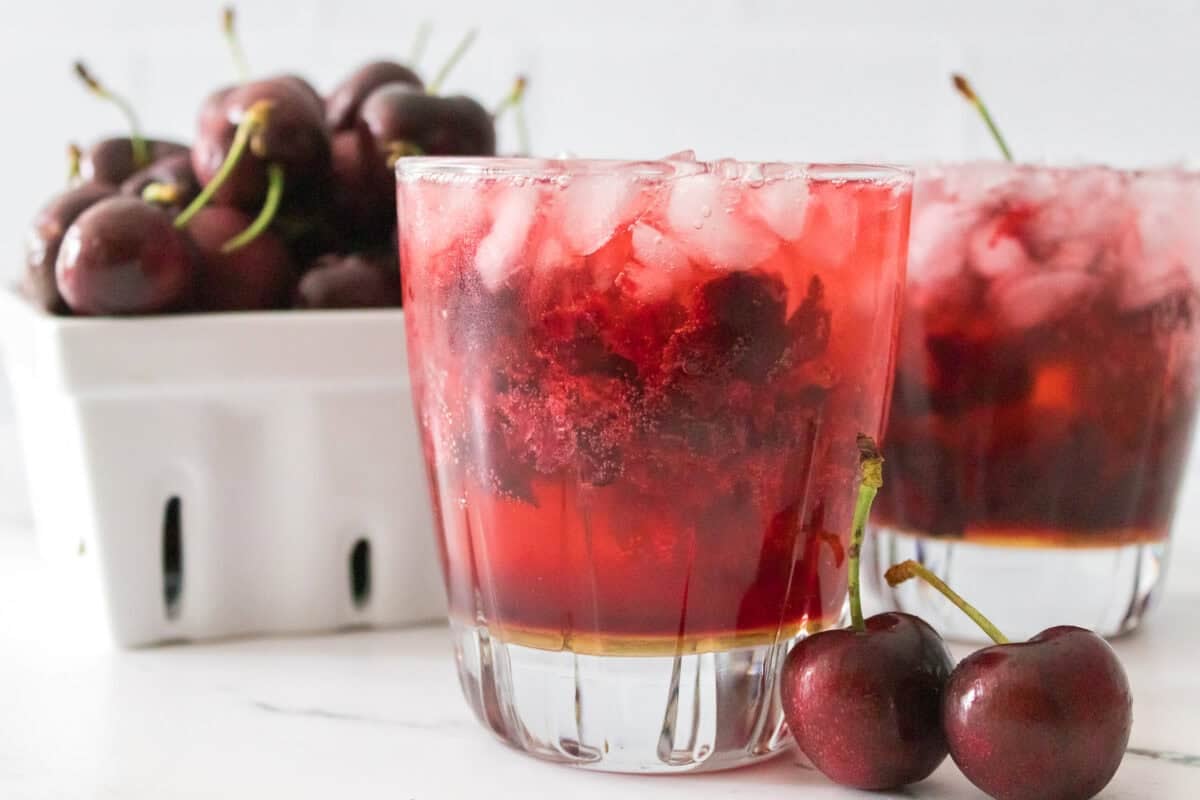 head on shot of cherry bomb cocktail in glass with bowl of cherries in background