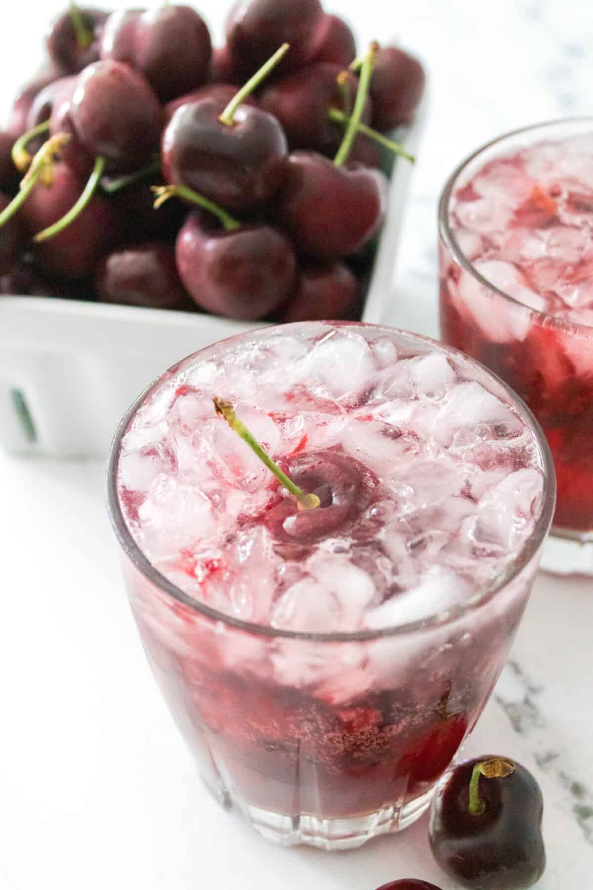 close up 45 degree shot of cherry bomb cocktail garnished with cherry with bowl of cherries in background