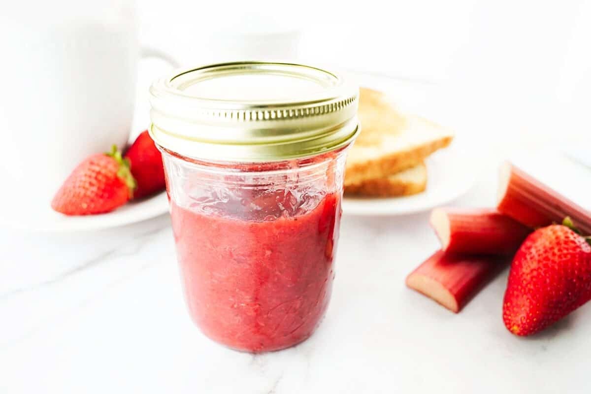 head on shot of strawberry rhubarb jam in mason jar with roast on plate in background with coffee