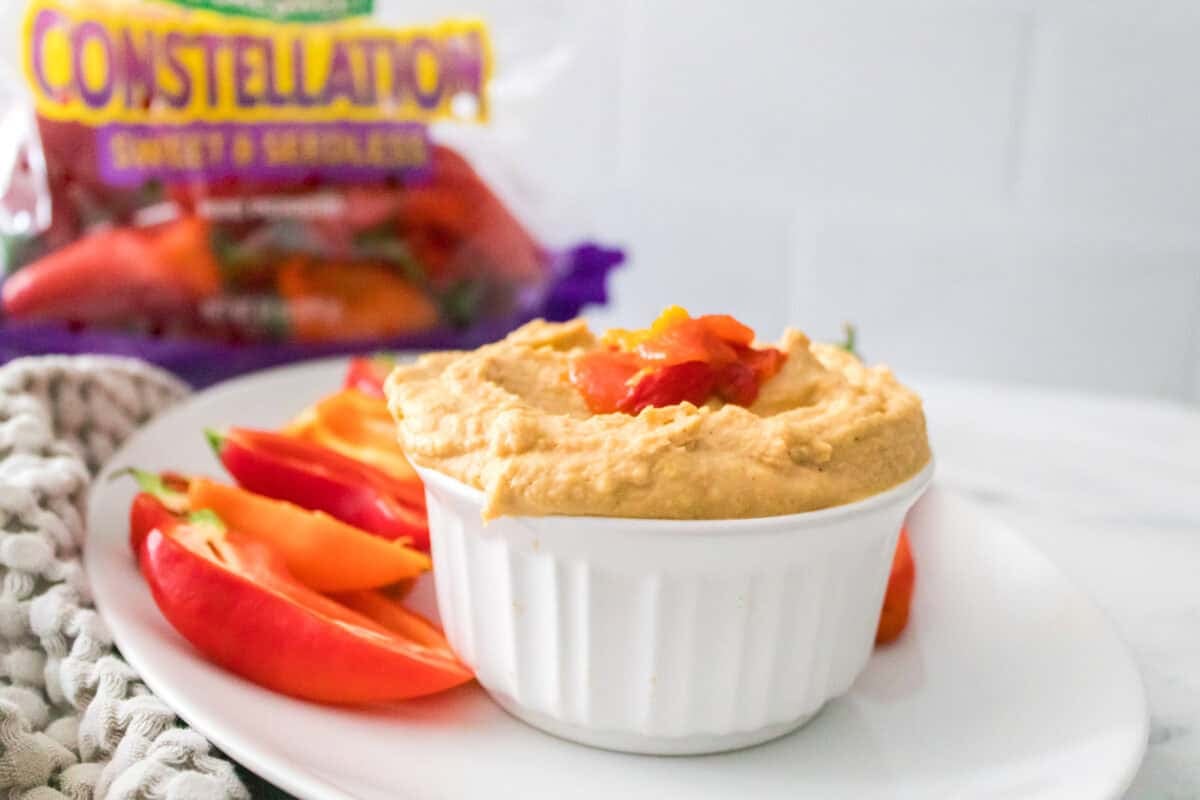 head on shot of roasted pepper hummus in white bowl served with sliced mini peppers with bag of naturesweet mini peppers in background