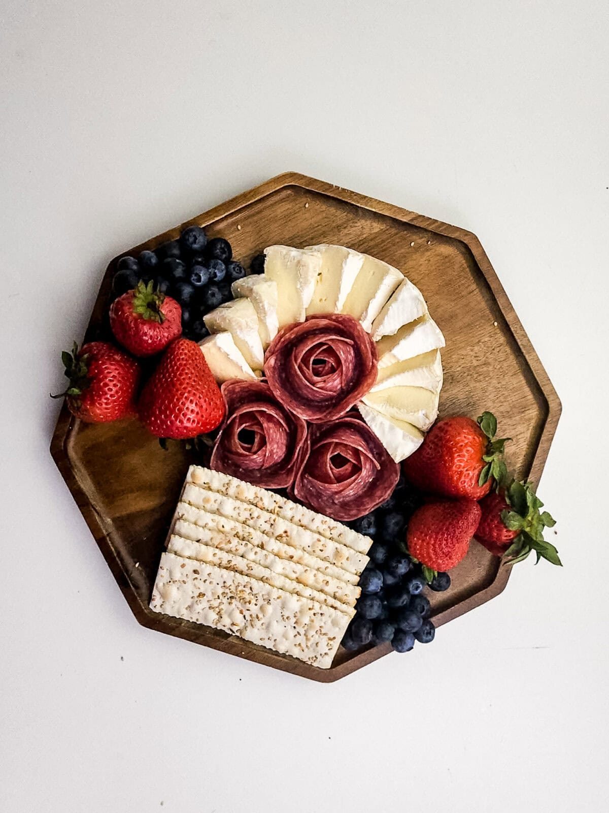 overhead shot of crackers, salami rosettes, brie cheese, blueberries and strawberries on cutting board
