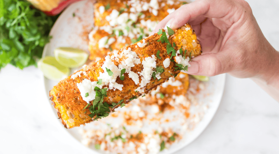 hand holding elote corn on the cob over white plate with more elote corn served with lime wedges