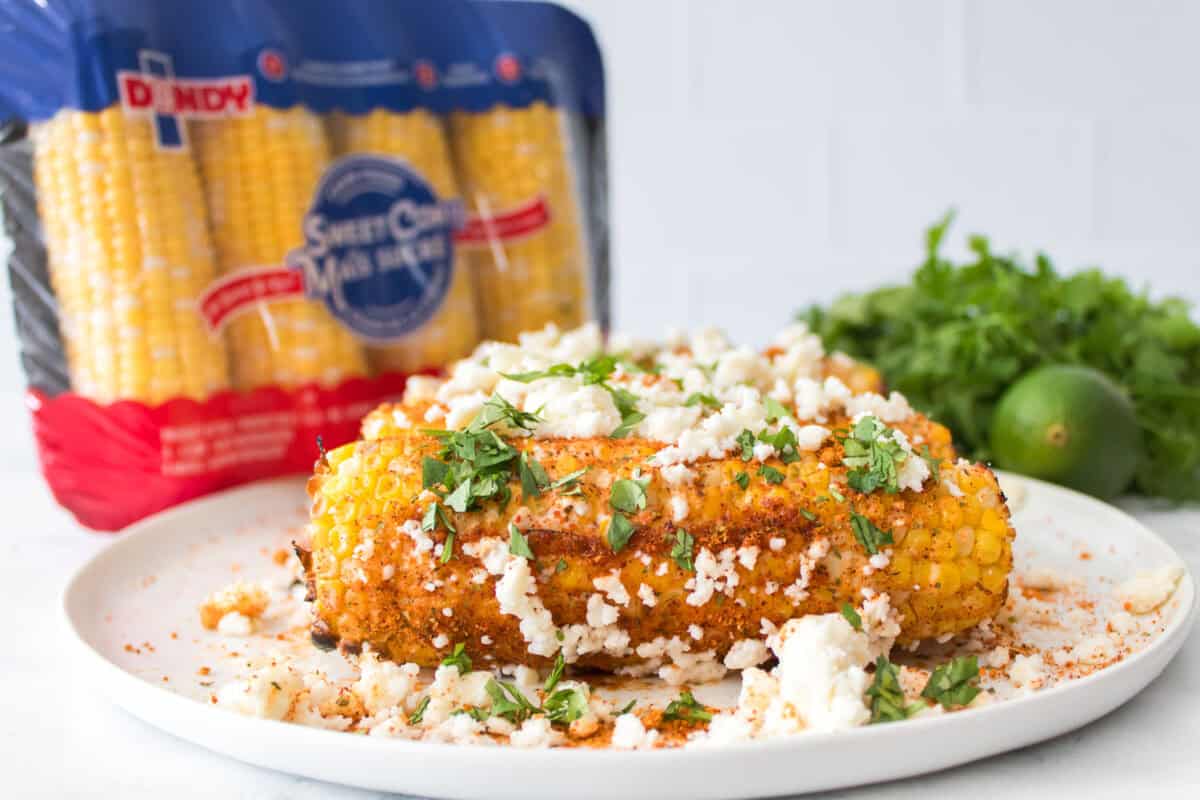 elote corn served on white plate with cilantro and lime in background with tray of dandy corn