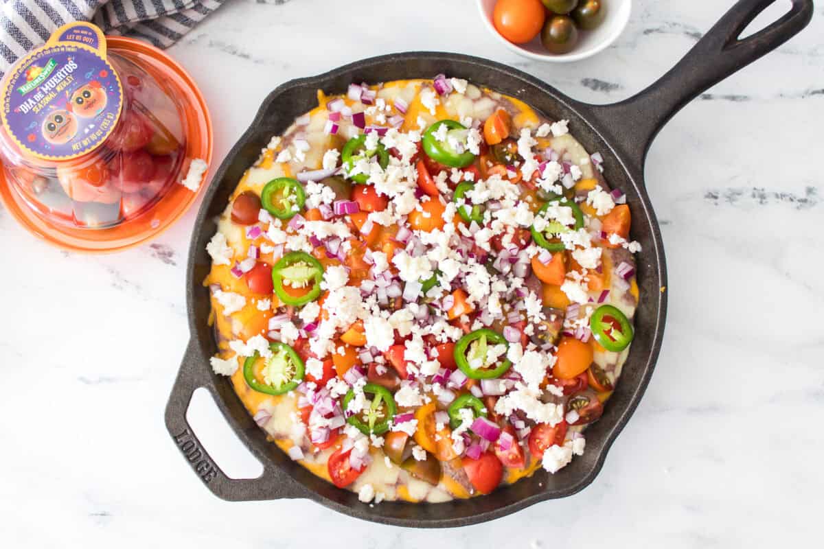 overhead shot of black bean dip in cast iron skillet topped with quartered cherry tomatoes, diced red onions, jalapeno slices and queso fresco cheese