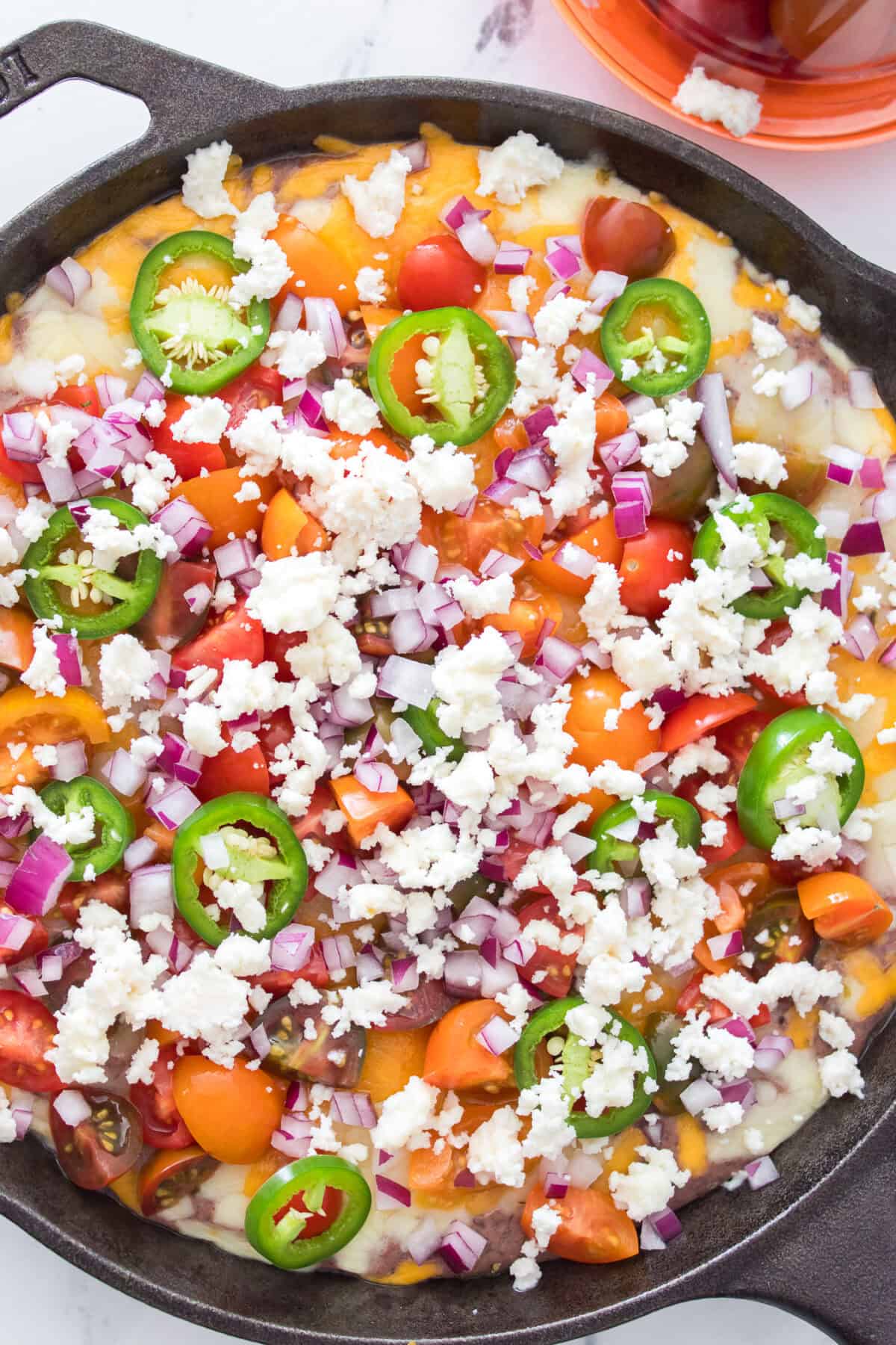 overhead close up shot of black bean dip in cast iron skillet topped with queso fresco, quartered cherry tomatoes, diced red onions and jalepenos