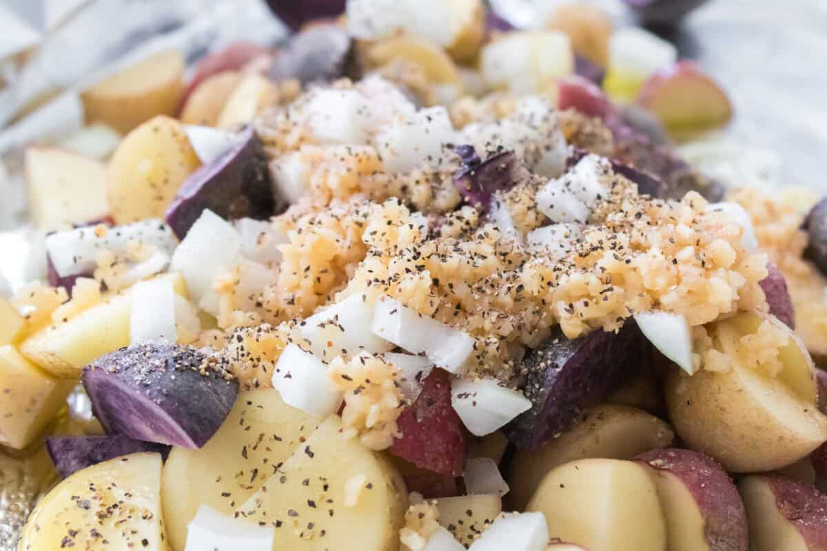 close up shot of quartered small medley potatoes, diced onion, minced garlic, salt, pepper and olive oil on baking sheet with aluminum foil before tossing together.