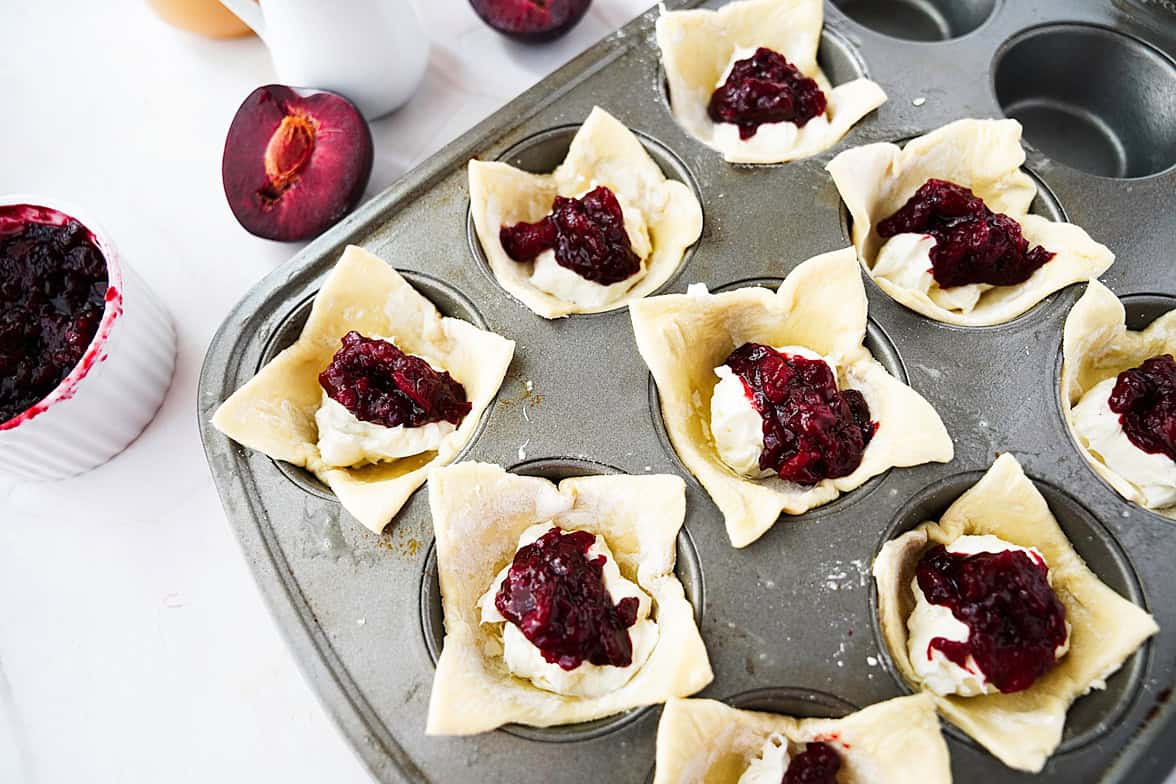 muffin tin with puff pastry that has been brushed with egg wash, with cream cheese mixture and plum jelly in each puff pastry previous to baking