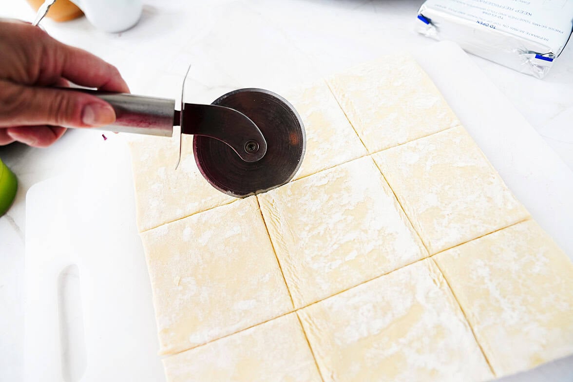 puff pastry on white cutting board with hand using pizza cutter to cut puff pastry into 9 pieces