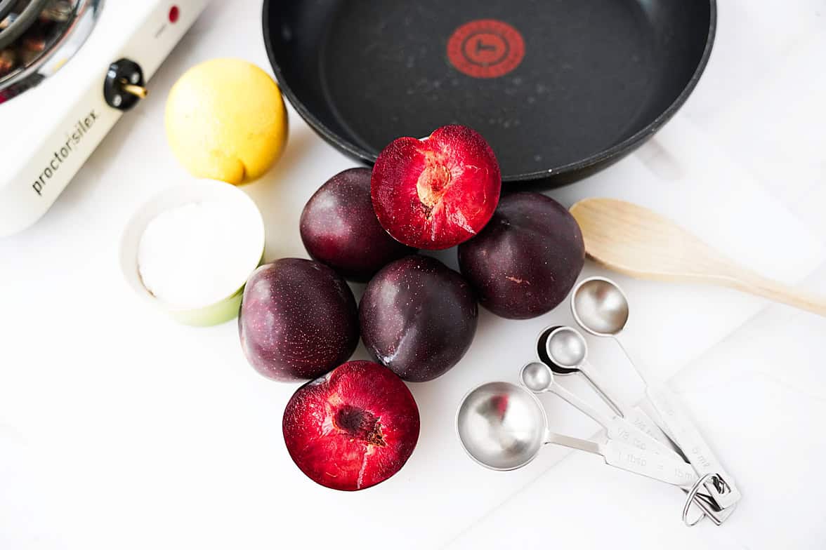 ingredients to make plum jelly recipe: plums, sugar, lemon, measuring spoons, wooden spoon, skillet