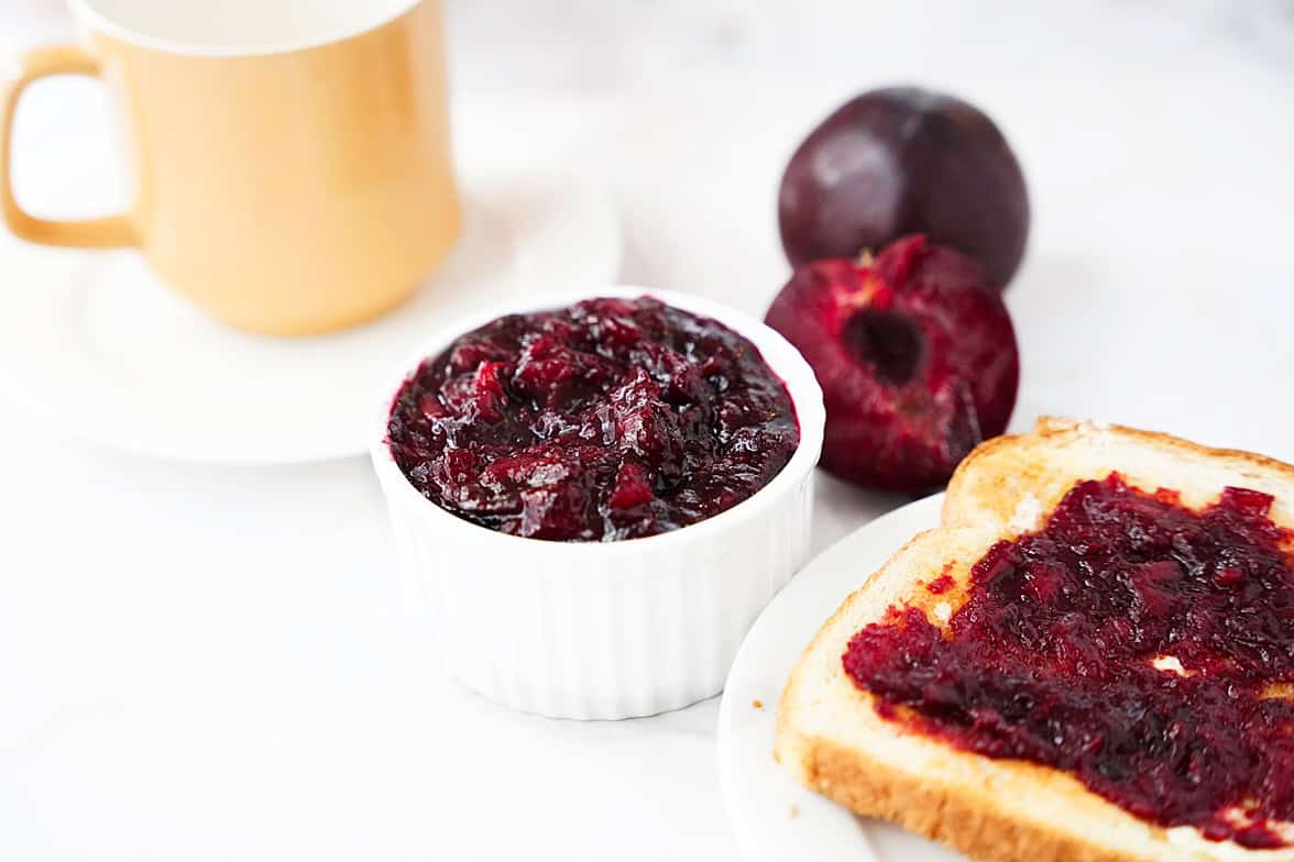 plum jelly in white ramekin with toast slathered with plum jelly on plate, halved plum in background with cup of coffee