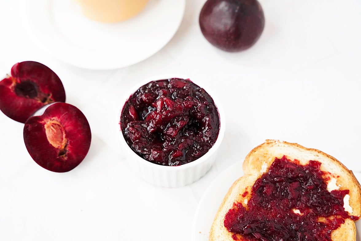 overhead shot of plum jelly in white ramekin with piece of toast in bottom right corner with butter and jelly, plum cut in half