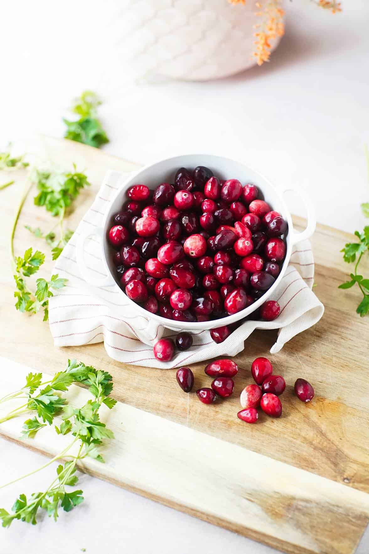 Vertical image of cranberries ina. white bowl on a cutting board with herbs