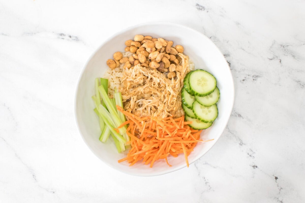 overhead shot of chicken and rice bowl put together white jasmine rice, topped with shredded chicken in peanut sauce, juelienned celery, shredded carrots, sliced cucumbers and peanuts